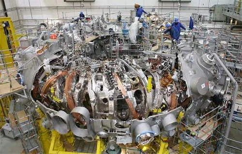 Reator de fusão nuclear em construção na Itália: estrutura toroidal metálica complexa com bobinas, fiações e operários em andaimes.