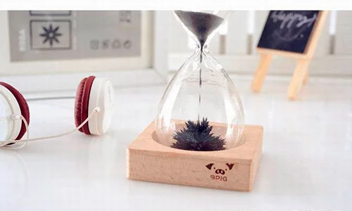 Ampulheta magnética de vidro, com areia de ferro preta caindo e formando uma escultura pontiaguda na base de madeira.