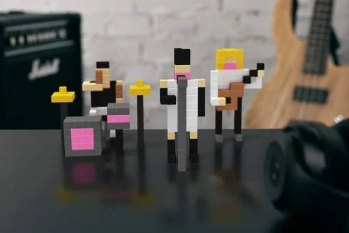 Avante lego blocos: Banda de bonecos pixelados com baterista, vocalista e guitarrista. Em mesa escura, com amp Marshall e guitarra ao fundo.
