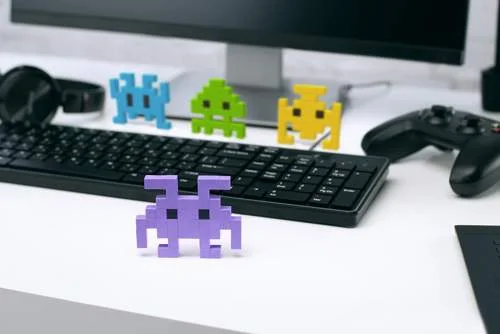 Avante: figuras de blocos coloridos pixel art (Space Invaders) em frente a teclado, monitor e controle de videogame.