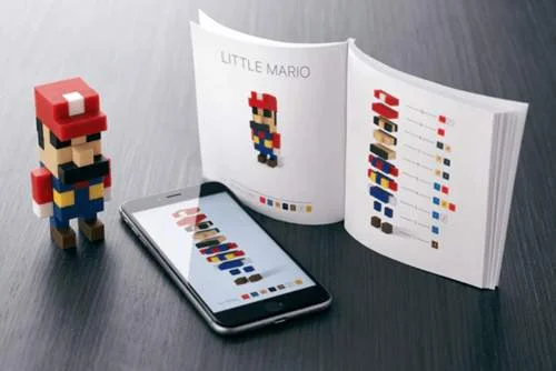 Boneco Mario pixelado feito de blocos LEGO, com smartphone exibindo suas peças e livro Little Mario aberto sobre mesa escura.