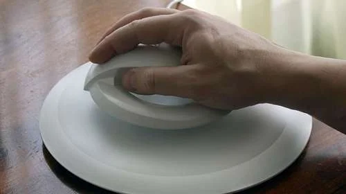 Mouse ergonômico bat flutuante branco, de design que evita dores, sendo segurado por mão sobre base circular.