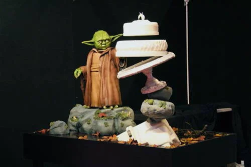 Bolo do Mestre Yoda levita pela força, com figura de Yoda em rochas e três andares de bolo branco flutuando ao lado.
