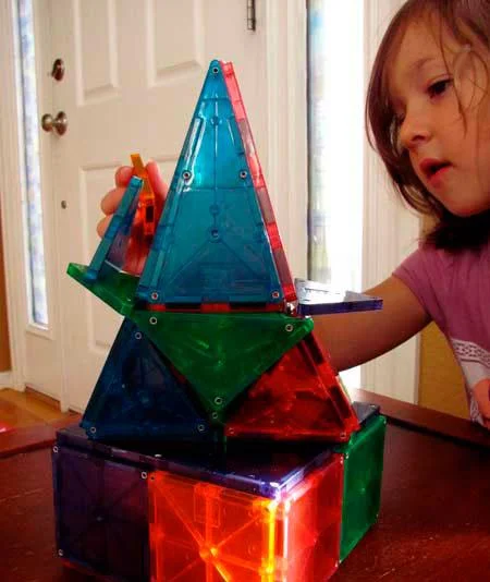 Brinquedo de ímãs 2. Estrutura colorida de peças magnéticas translúcidas triangulares e quadradas, sendo montada por uma criança.