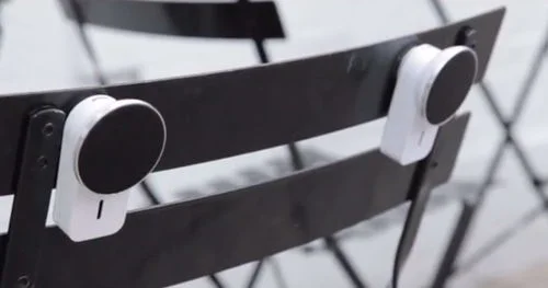 Duas caixas de som magnéticas brancas, com frente preta, fixadas nos encostos de uma cadeira preta de metal.