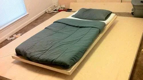 Cama flutuante com colchão branco, edredom e travesseiro verde em plataforma de madeira clara. Quarto com janela ao fundo.