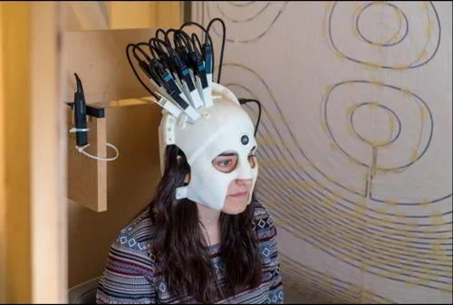Capacete branco com múltiplos cabos na cabeça de uma mulher. Este capacete auxilia na realização de exames no cérebro.