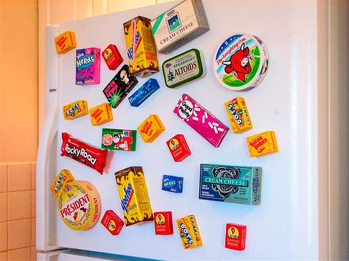 Imãs de geladeira de miniaturas coloridas de doces e alimentos diversos colecionados na porta branca de um refrigerador.