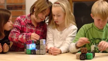 Cubelets, os robôs modulares, sendo montados por quatro crianças. Peças cúbicas coloridas, uma delas com luz azul, sobre mesa de madeira.