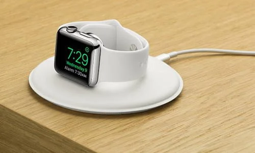 Dock magnética para Apple Watch 2, prata com pulseira branca, exibindo 7:29, sobre base de carregamento branca em mesa de madeira.