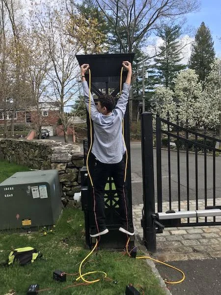 Eletroímãs e os poderes do Homem-Aranha: Jovem com fios amarelos e caixas pretas nos pés e mãos, agarrado a um poste de portão.