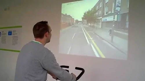 Exercitando com o Street View: homem em bicicleta ergométrica e rua projetada na parede.