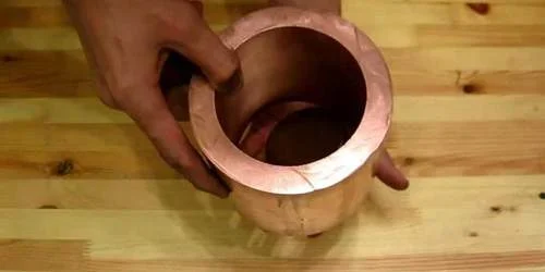 Tubo de cobre e imã de neodímio: Mãos seguram tubo de cobre, com imã escuro parcialmente dentro, para experiência em mesa de madeira.