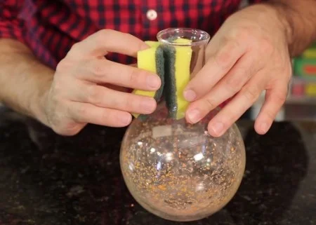 Mãos inserem esponja amarela em balão de vidro com partículas para criar uma esponja magnética em experimento com imãs.