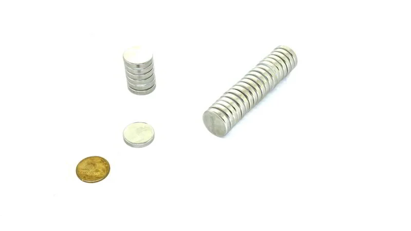 Ímãs de neodímio 25x5mm prateados, circulares, empilhados, em fila e soltos, com moeda dourada para escala.