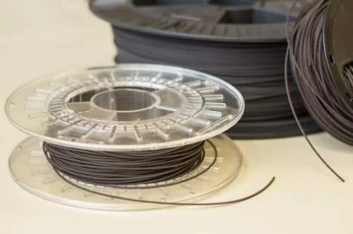 Rolo de filamento preto para impressão 3D em carretel transparente, com outro rolo escuro ao fundo.