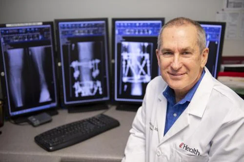 Médico sorridente em jaleco branco diante de 4 monitores exibindo imagens de raios-X de membros e fixadores.