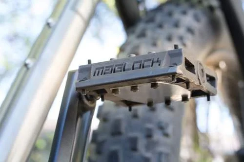 Pedal magnético MAGLOCK para bicicleta. Retangular, cinza, com pinos de aderência e o logo MAGLOCK. Montado na bike, pneu ao fundo.