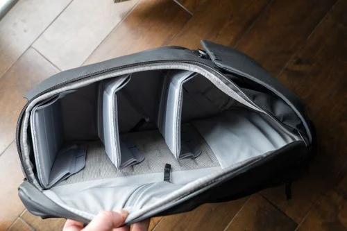 Mochila preta com fecho magnético, aberta, exibindo três divisórias internas acolchoadas cinzas.