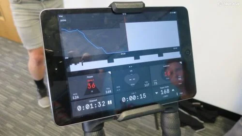 Wattbike Atom com tablet exibindo precisão de treinamento profissional: gráficos de potência, velocidade e frequência cardíaca.