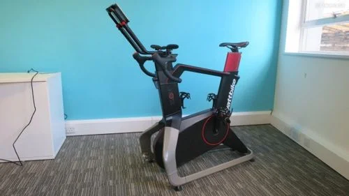 Wattbike Atom, bicicleta ergométrica preta e vermelha de design robusto, sobre carpete cinza e fundo de parede azul clara.