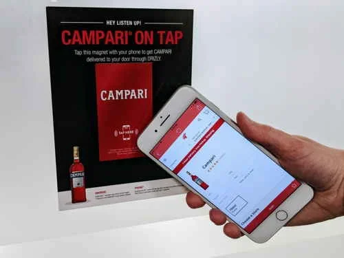 Display Campari NFC: mão usa smartphone com app de entrega ao tocar imã para pedido de bebidas. Magneto em painel preto.