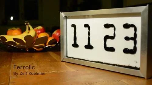 Relógio de ferrofluido em moldura prata, exibindo 1123 em preto sobre fundo branco. Fruteira com bananas e maçãs ao fundo.