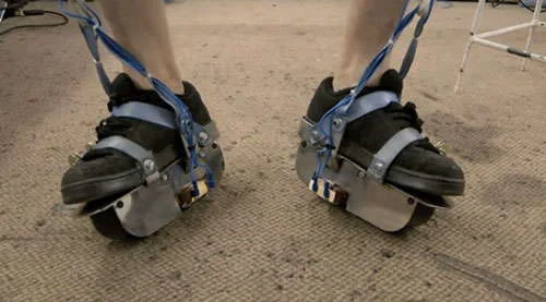 Sapatos magnéticos, a nova criação de Colin Furze. Tênis pretos com plataformas metálicas e correias azuis, sobre carpete.