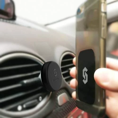 Suporte magnético para smartphones, disco preto na ventilação do carro. Mão posiciona smartphone com placa magnética retangular e logo S.