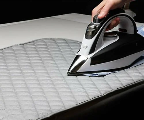 Tábua de passar roupa magnética cinza acolchoada, com ferro de passar roupa preto e branco sendo usado por uma mão.