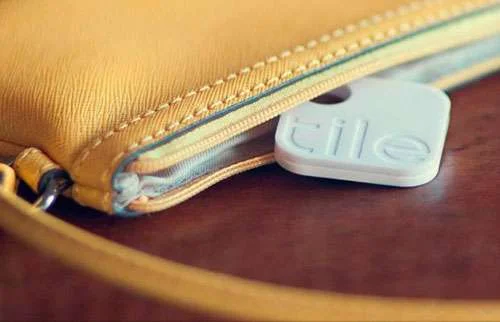 Tile, dispositivo que localiza objetos perdidos, branco e retangular, saindo de uma carteira amarela com zíper sobre mesa de madeira.