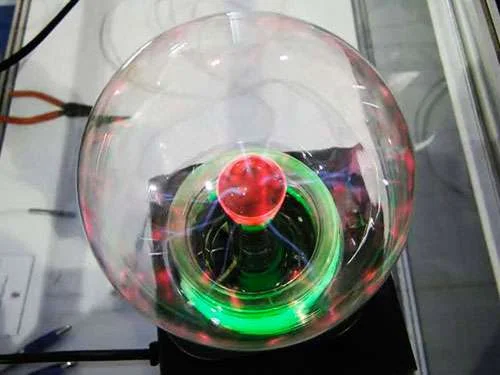 Globo de plasma transparente com esfera central vermelha, anel verde luminoso e filamentos elétricos azuis sobre base preta.